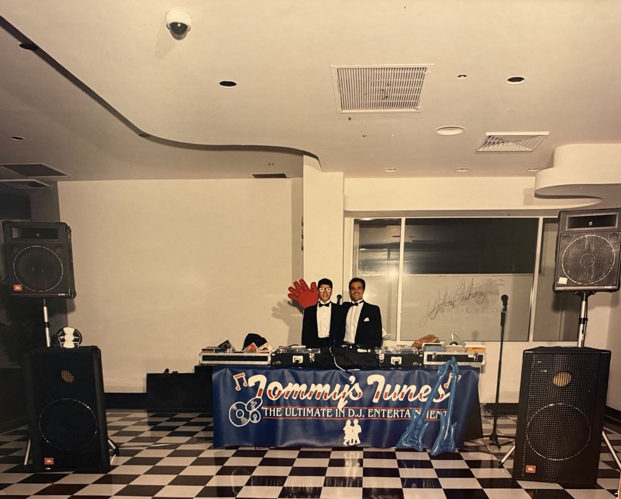 A Tommy's Tunes DJ setup from the early years: two DJs in tuxedos behind a banner, JBL speakers either side, checkerboard floor